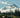 Snow-capped mountain peak with cloud cover, dense evergreen forest, and wildflowers in the foreground.