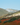 Rolling hills with misty slopes, vegetation, and a wooden fence under a clear blue sky.